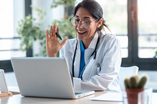 Female-doctor-waving-and-talking-with-colleagues-through-a-video-call-with-a-laptop-in-the-consultation.-1249601252_6218x4145